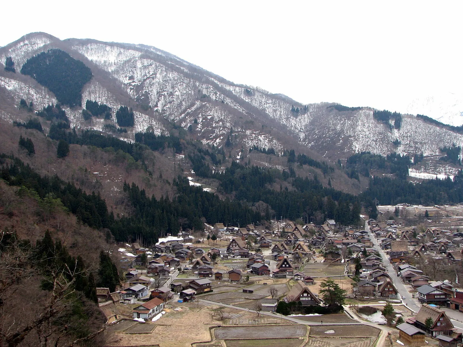 The village of Shirakawa go seen from the viewpoint above it, thatched roofs below snow covered mountains
